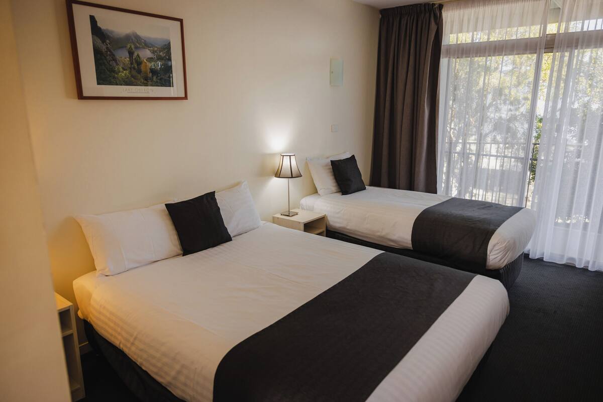A hotel room with two beds, a nightstand, a lamp, a framed picture on the wall, and a window with curtains.