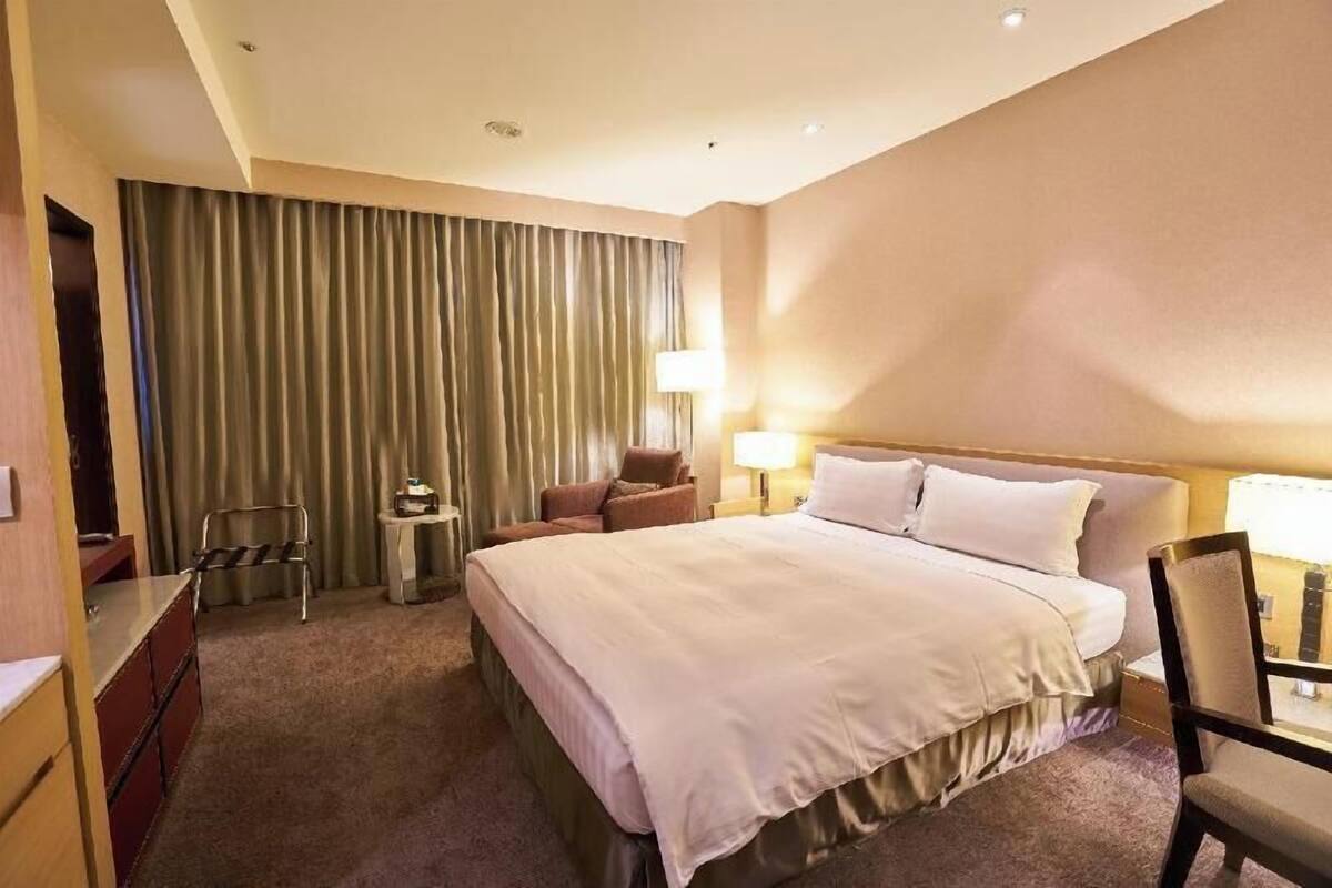 Quarto de hotel com uma cama grande, duas mesas de cabeceira, uma escrivaninha, uma cadeira, uma luminária e uma cortina.