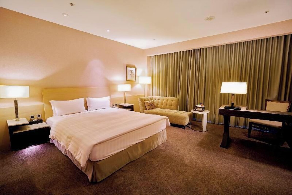 Habitación de hotel con una cama grande, dos mesitas de noche con lámparas, un pequeño sofá, un escritorio con una silla y un ventanal con cortinas.