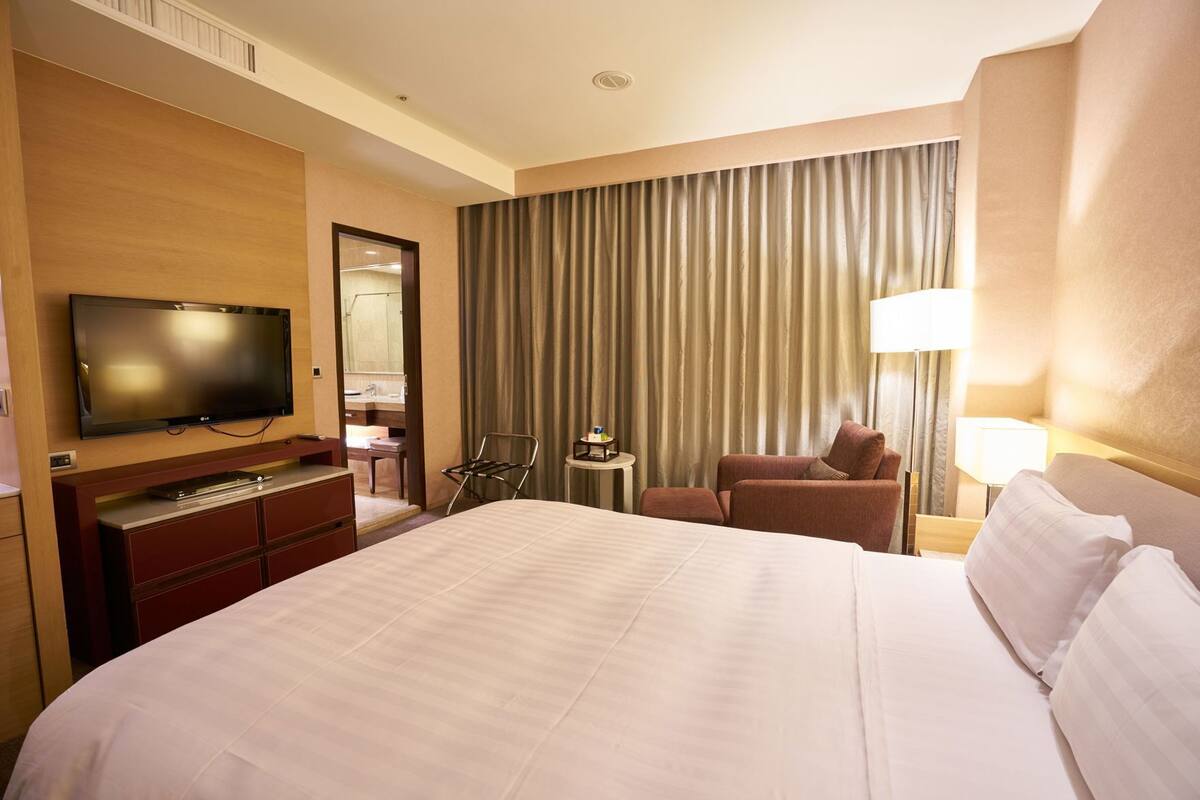 Quarto de hotel com uma cama grande, televisão, escrivaninha com cadeira, mesa pequena e janela com cortinas.