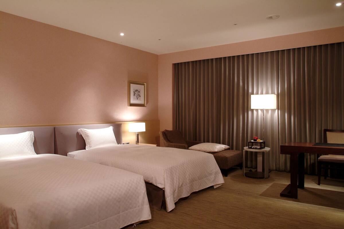 Quarto de hotel com duas camas, uma cadeira, uma escrivaninha, um abajur e uma janela grande com cortinas.