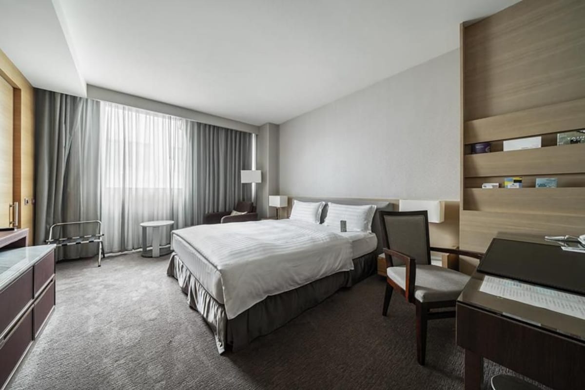 Una habitación de hotel con cama, escritorio, silla, lámpara y estantería.