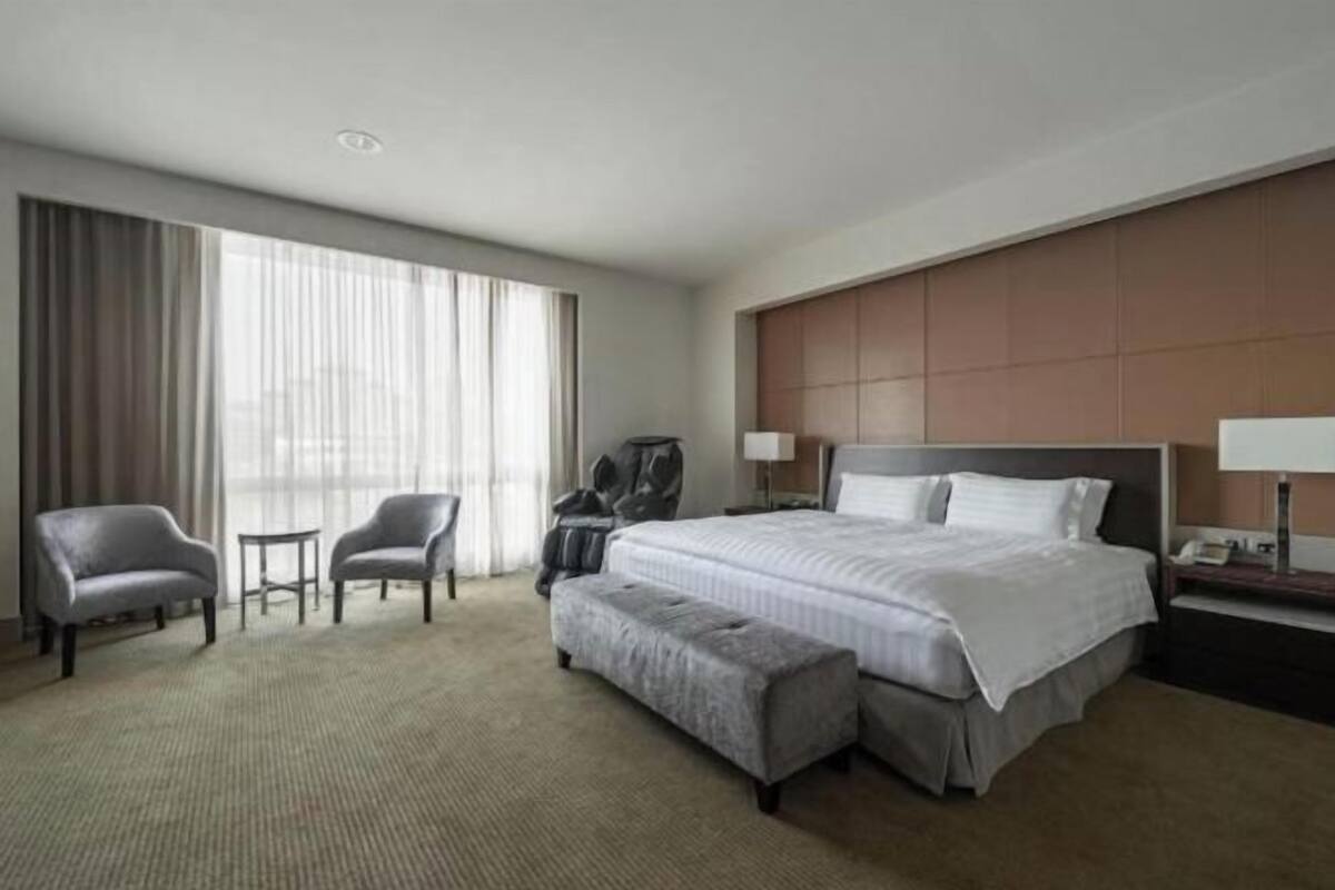 Quarto de hotel com uma cama grande, duas poltronas, uma mesinha, um banco, um guarda-roupa e uma mesa de cabeceira com uma luminária.