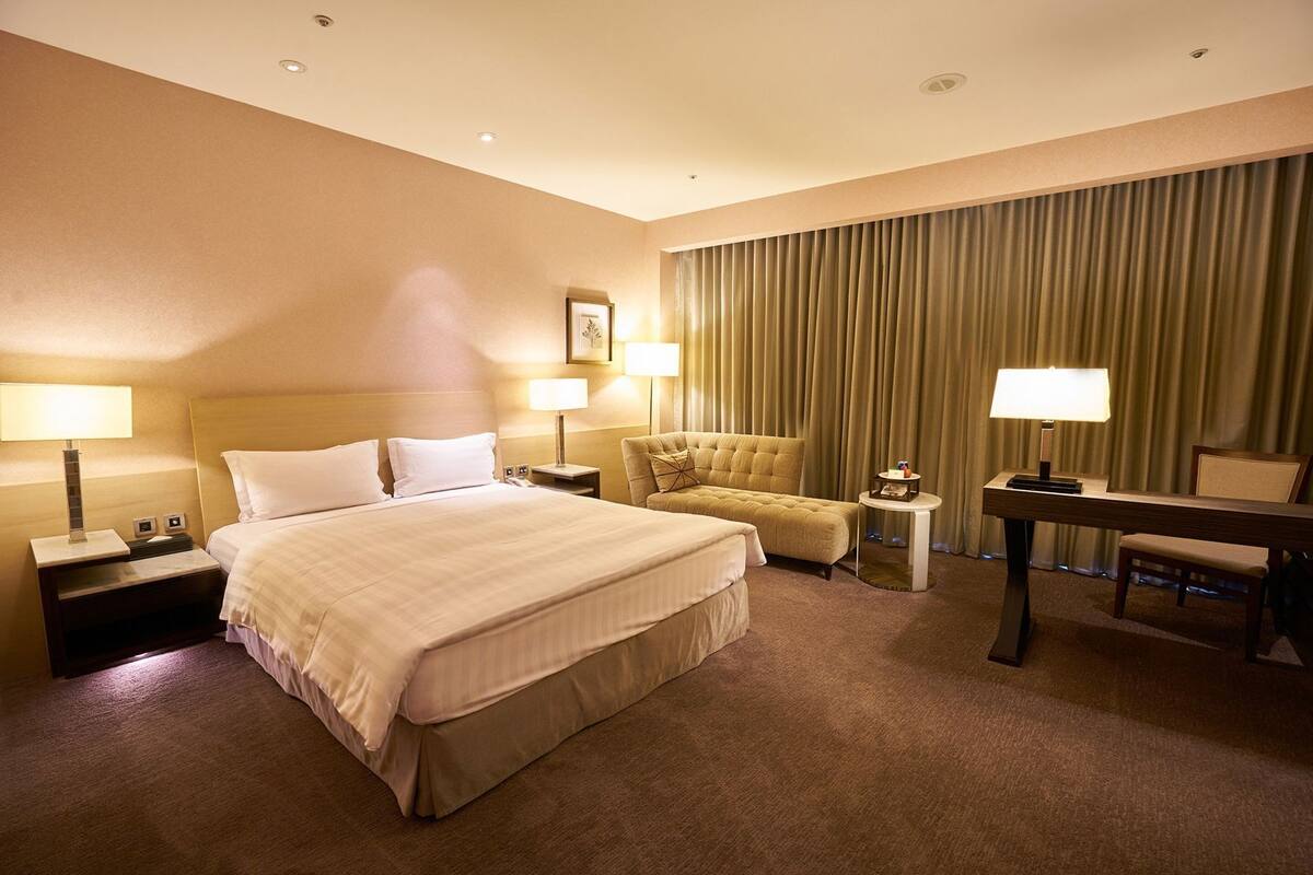 Quarto de hotel com uma cama grande, duas mesas de cabeceira, uma escrivaninha, uma cadeira e uma janela ampla com cortinas.