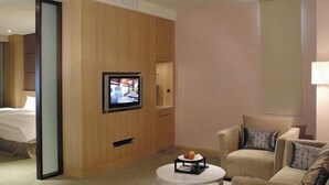 A hotel room with a bed, a TV mounted on a wooden panel, a round coffee table with a fruit bowl, and two armchairs.
