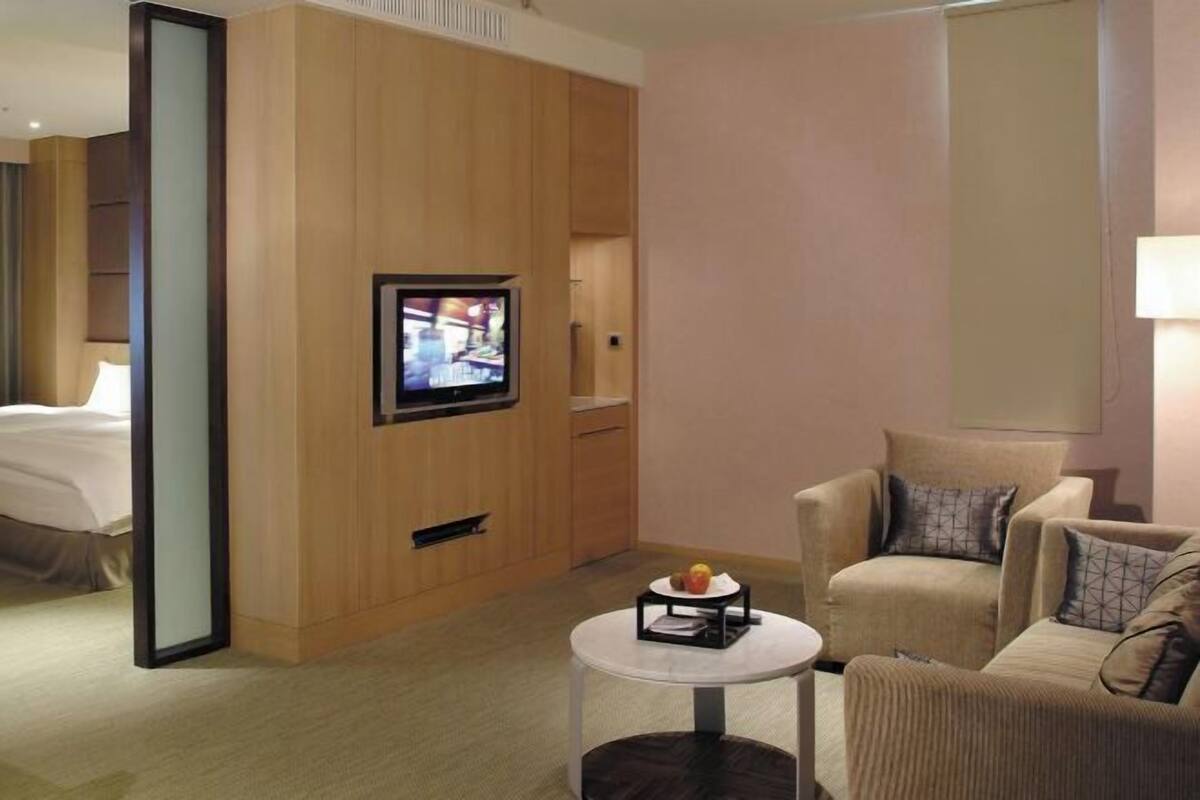 Quarto de hotel com uma cama, uma televisão fixada numa estrutura de madeira, uma mesa de centro redonda com uma fruteira e duas poltronas.