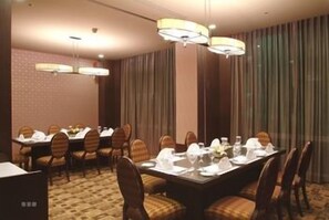 En hotels spisestue med et langt bord dækket til et måltid, omgivet af stole, og udsigt til byen gennem store vinduer.