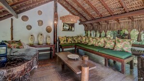 A cozy lounge area with a thatched roof, a green sofa with floral cushions, a wooden coffee table, and decorative vases on the wall. - Pousada Villa N'Kara (Itacaré)