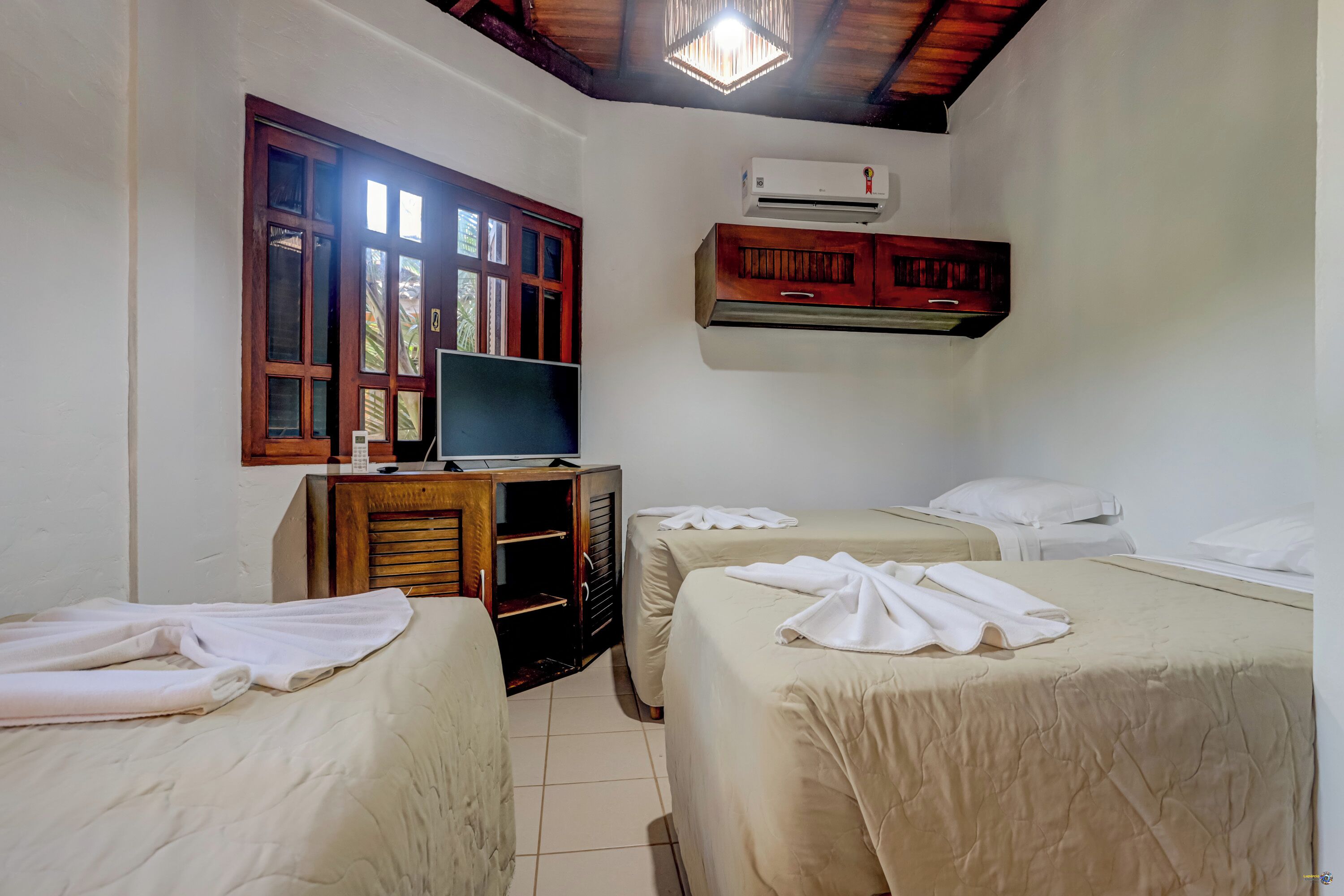 A hotel room with two beds, a TV on a wooden cabinet, an air conditioner, and a window with wooden shutters.