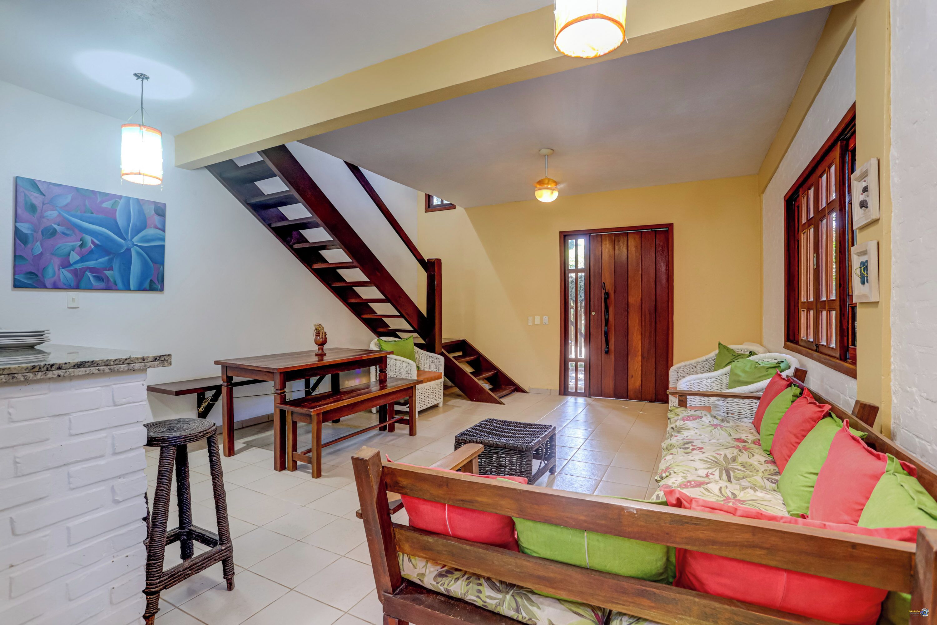 Amplia sala de estar con una escalera de madera, un sofá con cojines de colores, una mesa de comedor y una pared de ladrillo blanco.