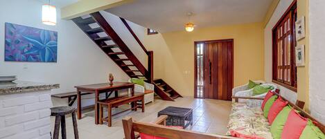 A spacious living area with a wooden staircase, a sofa with colorful cushions, a dining table, and a white brick wall.