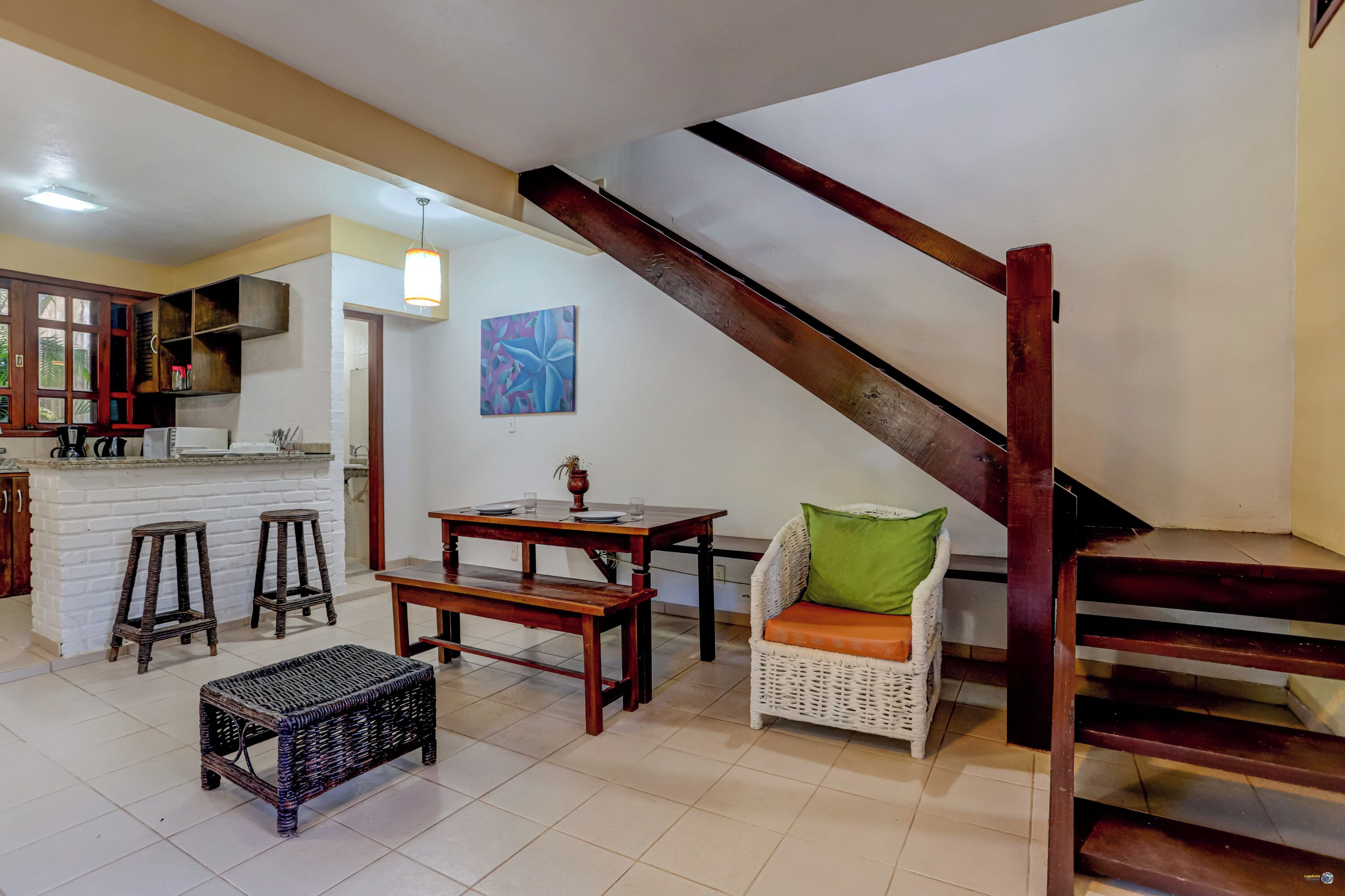 A kitchen with a white island, bar stools, and a dining table with chairs. There is a wicker bench and a wicker basket. A staircase with wooden railings is visible.