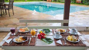 A breakfast setup with various dishes and beverages on a table, overlooking a swimming pool. - Pousada Villa N'Kara (Itacaré)