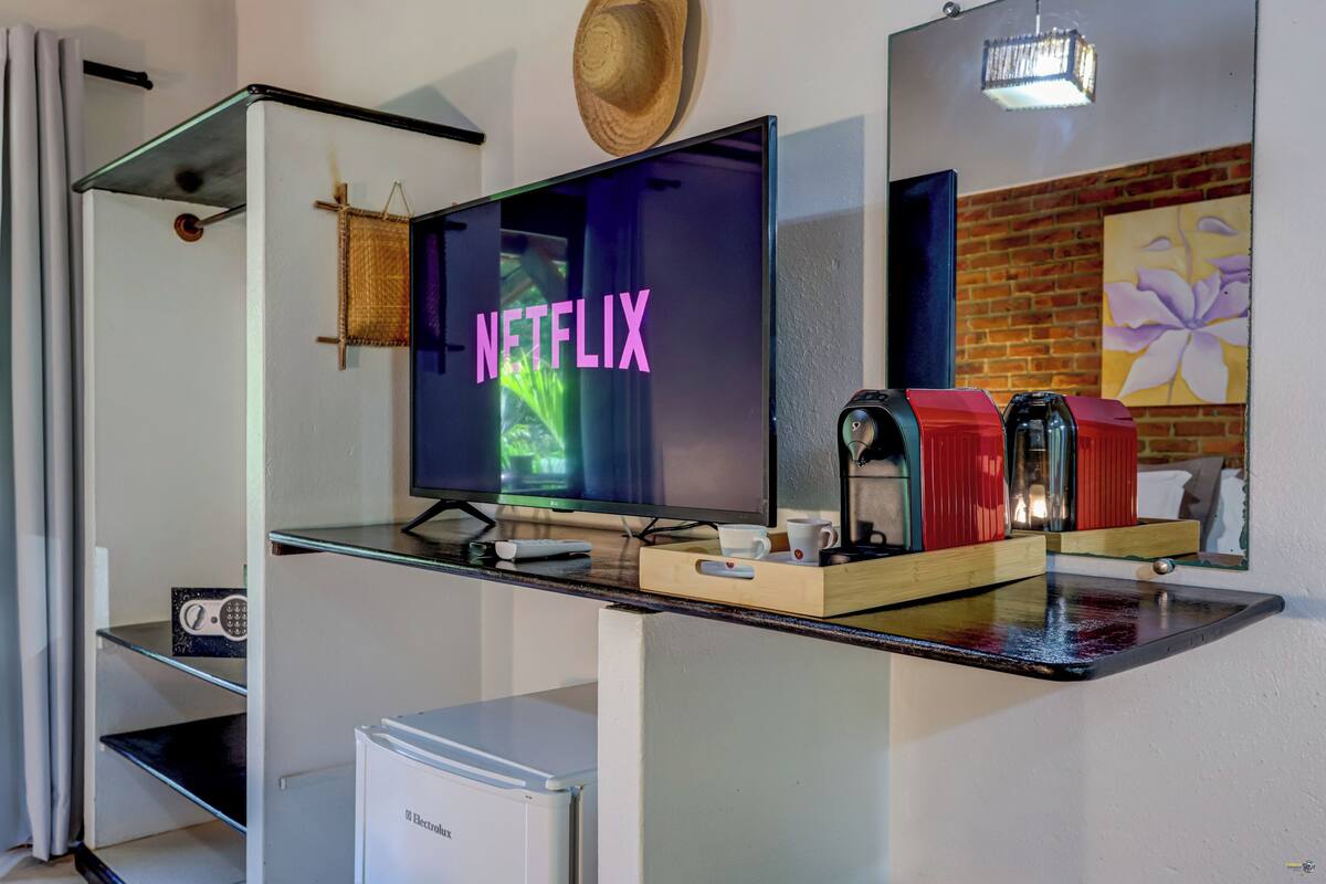 Habitación moderna con una mesa de centro de vidrio, un televisor de pantalla plana mostrando 'NETFLIX', un refrigerador pequeño y una pared de ladrillo con un cuadro de flores.