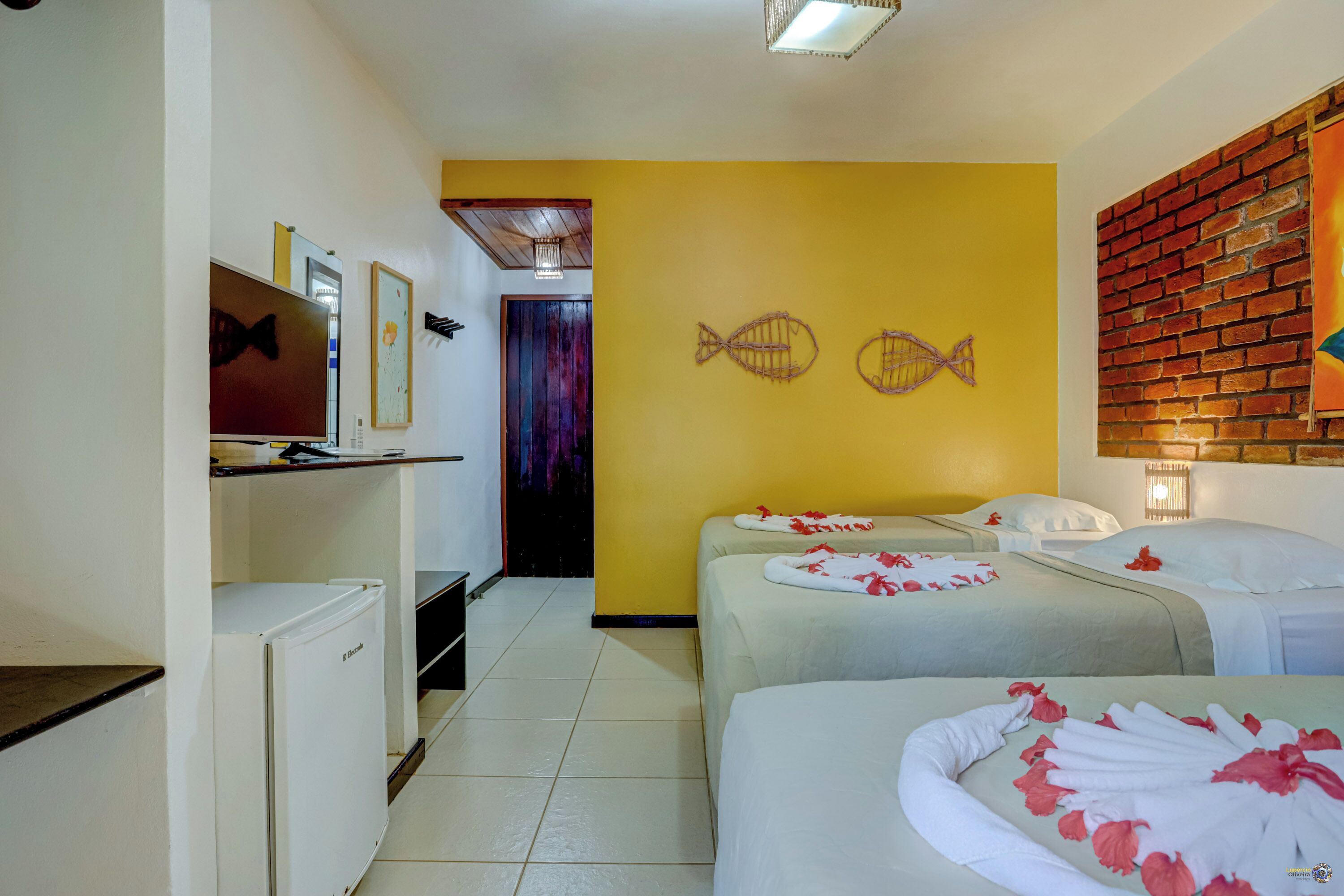 A hotel room with two beds, a television, a refrigerator, and a yellow wall with fish decorations.