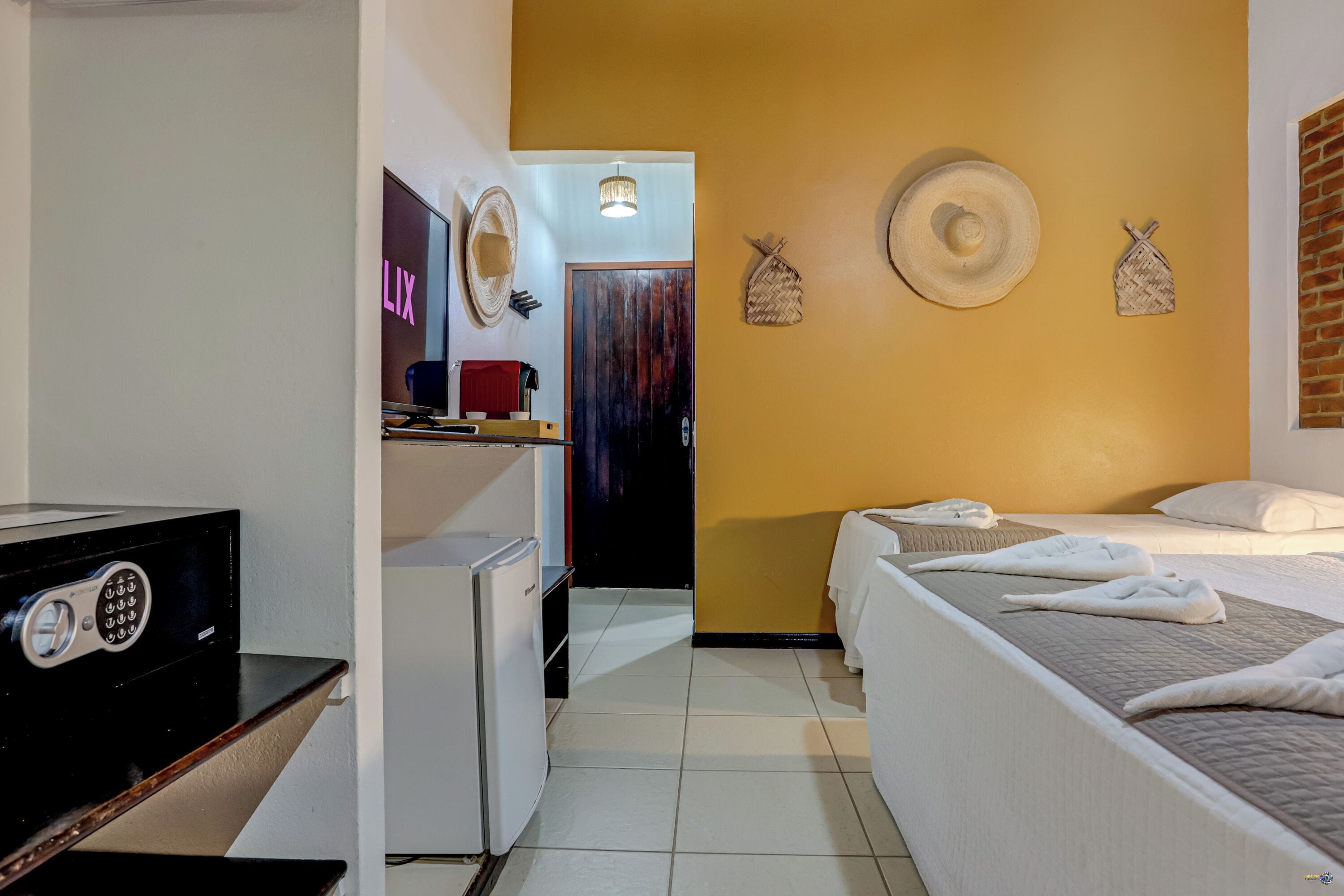 Habitación de hotel con cama, escritorio con computadora, refrigerador pequeño y televisor de pared mostrando 'LIX'.