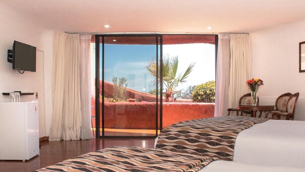 Habitación de hotel con cama, escritorio con silla, televisor montado en la pared y puerta corrediza de vidrio que da a un balcón con vistas al paisaje.