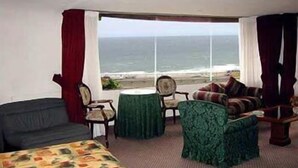 Una habitación con una cama, dos sillas, una mesita y un sofá. Hay un ventanal con vista al océano.