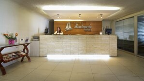 Zona de recepción de un hotel con un mostrador de madera, una recepcionista y suelo de baldosas.
