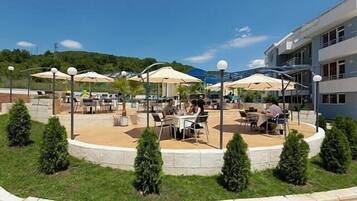 An outdoor dining area with round tables and umbrellas, surrounded by a low stone wall and small trees.