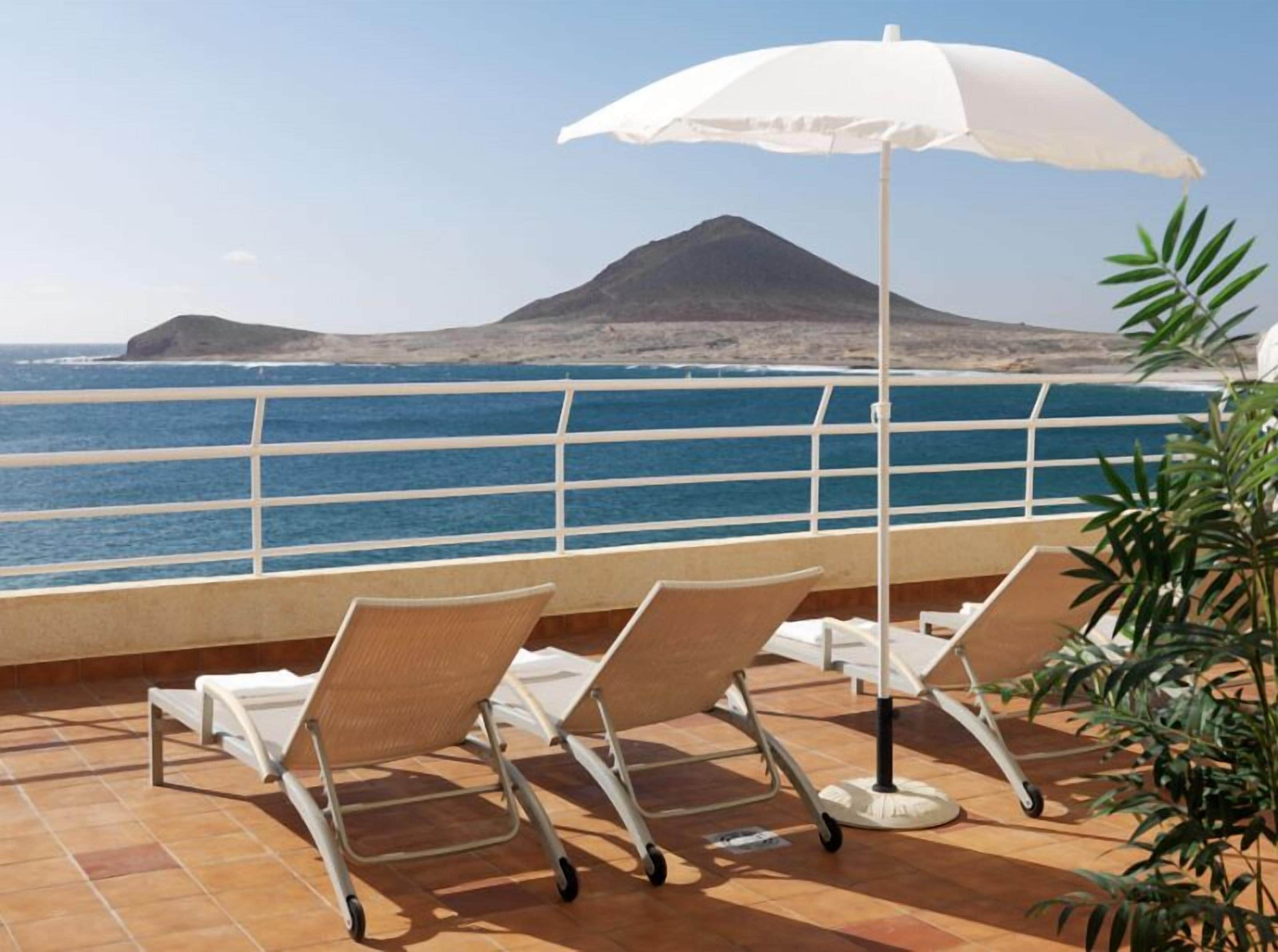 Una terraza con dos tumbonas y una sombrilla blanca, con vistas a la costa y una montaña al fondo.