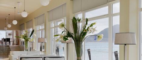 A hotel lobby with a long corridor, white furniture, large windows with a view of the sea, and decorative plants.