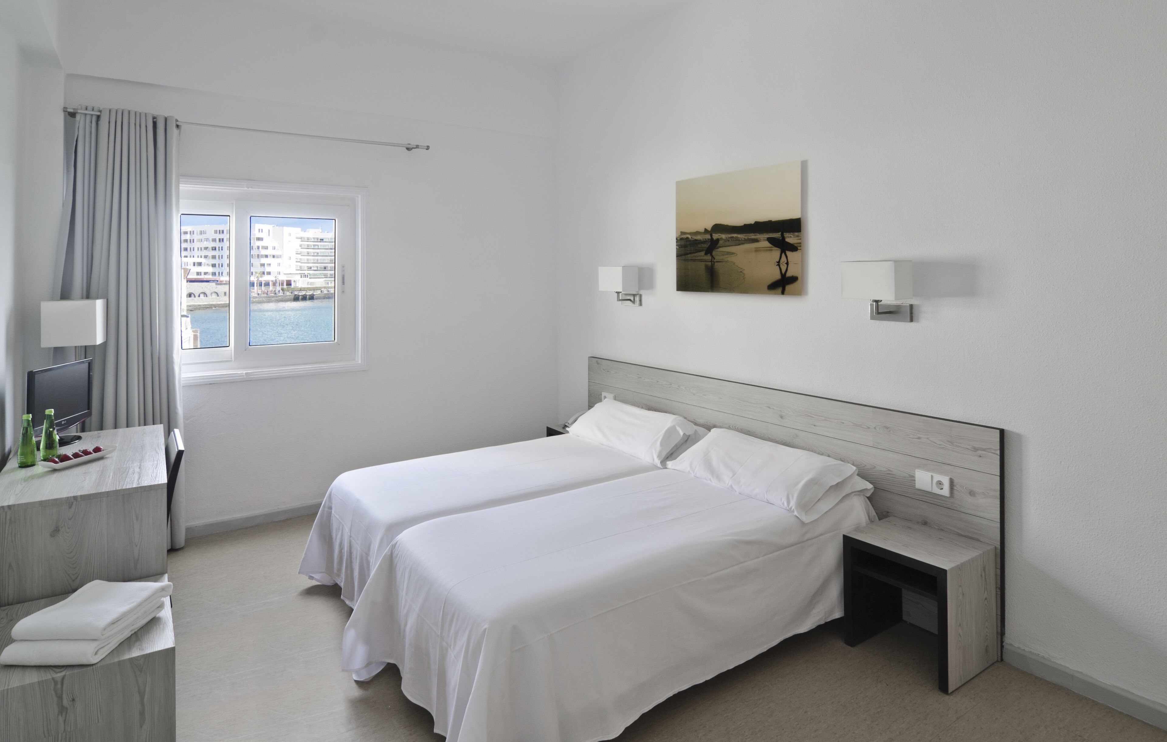 Quarto de hotel com duas camas, uma mesa com abajur, janela com vista para edifícios e um quadro na parede.