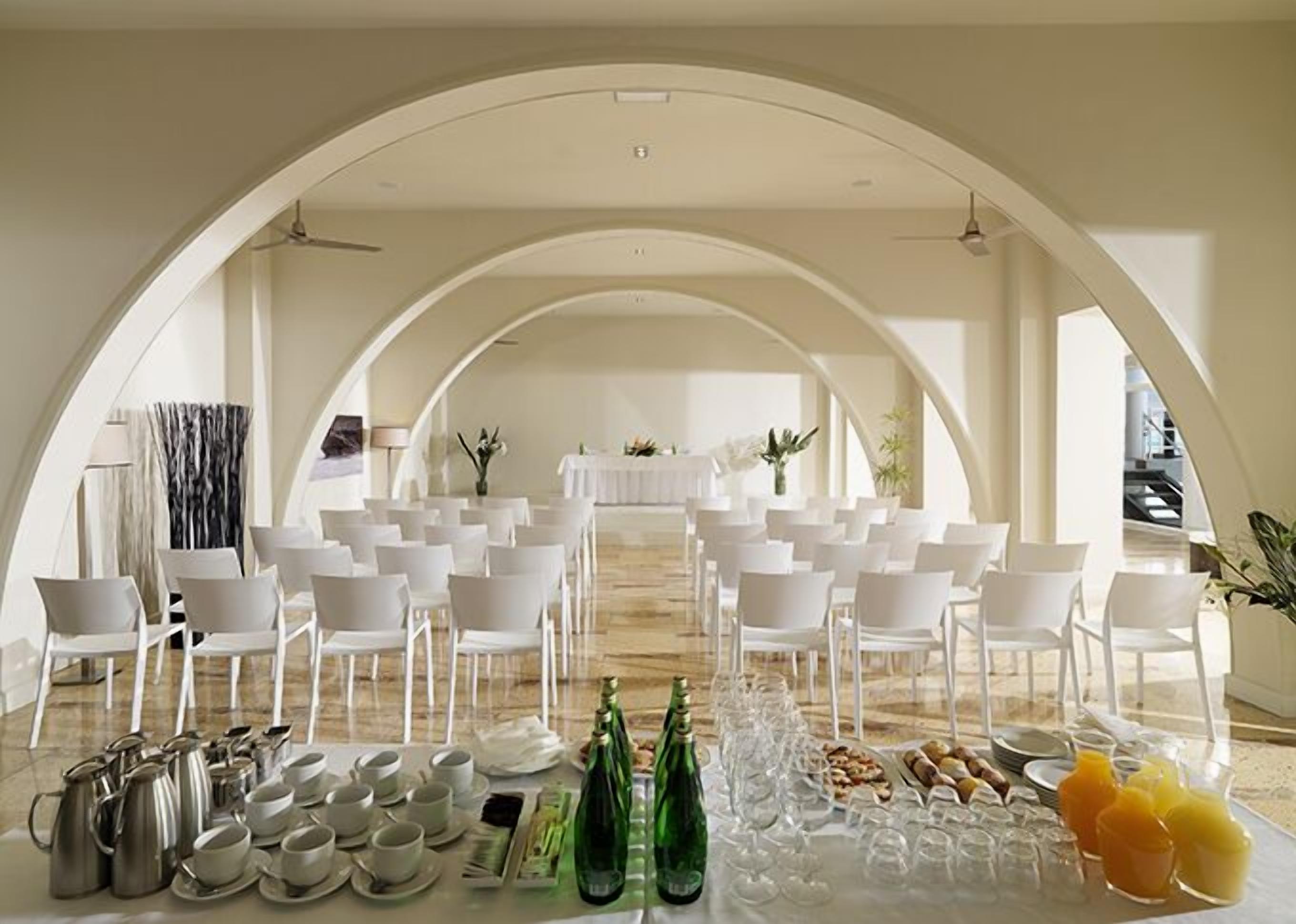 A formal dining setup with white chairs and tables, arranged in an arched room with a view of a balcony.