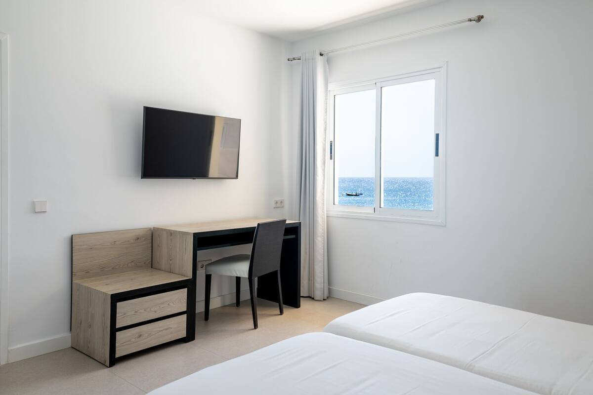 Ein Schlafzimmer mit Bett, Schreibtisch mit Stuhl, Fernseher und einem Fenster mit Meerblick.