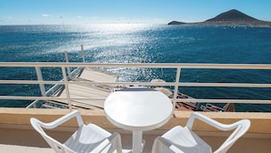 Un balcon avec une table blanche et deux chaises blanches, donnant sur une étendue d’eau, avec une montagne visible au loin.