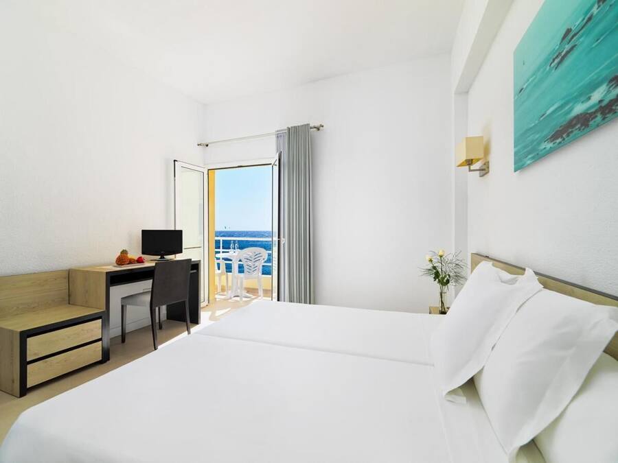 A hotel room with a bed, a desk with a computer, a chair, and a view of the sea through a large window.