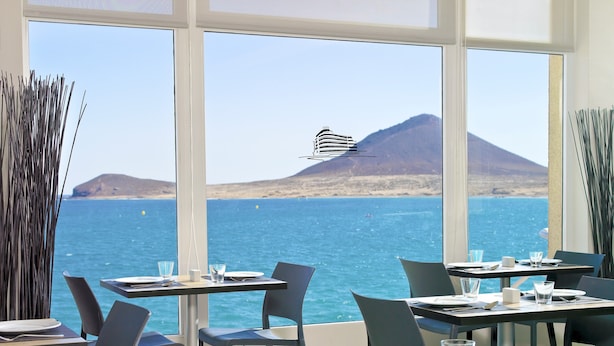 Un espace repas avec des tables et des chaises, de grandes fenêtres offrant une vue sur la mer et une montagne.
