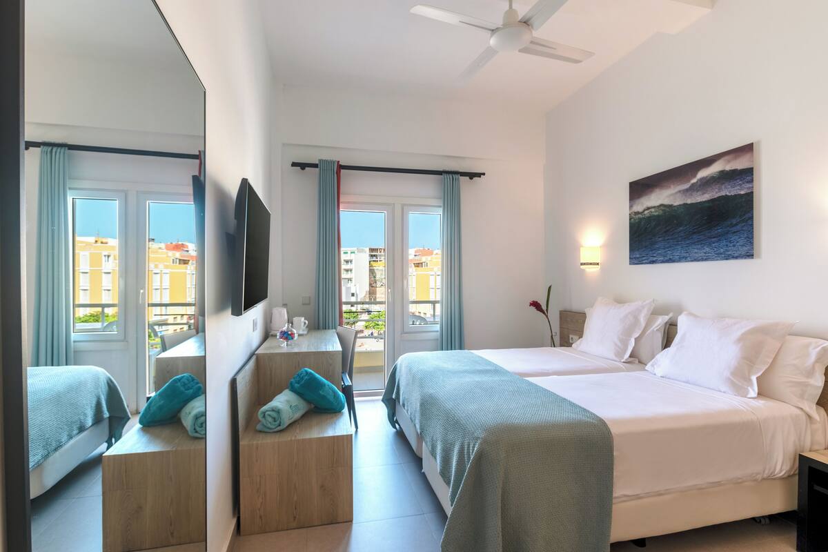 Ein Hotelzimmer mit zwei Betten, einem Schreibtisch, einem Stuhl, einem Fernseher und einem Balkon mit Vorhängen.