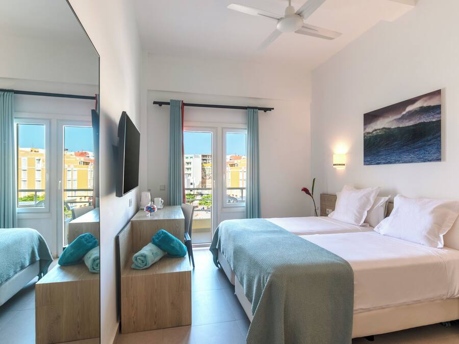 Ein Hotelzimmer mit zwei Betten, einem Schreibtisch, einem Stuhl, einem Fernseher und einem Balkon mit Vorhängen.