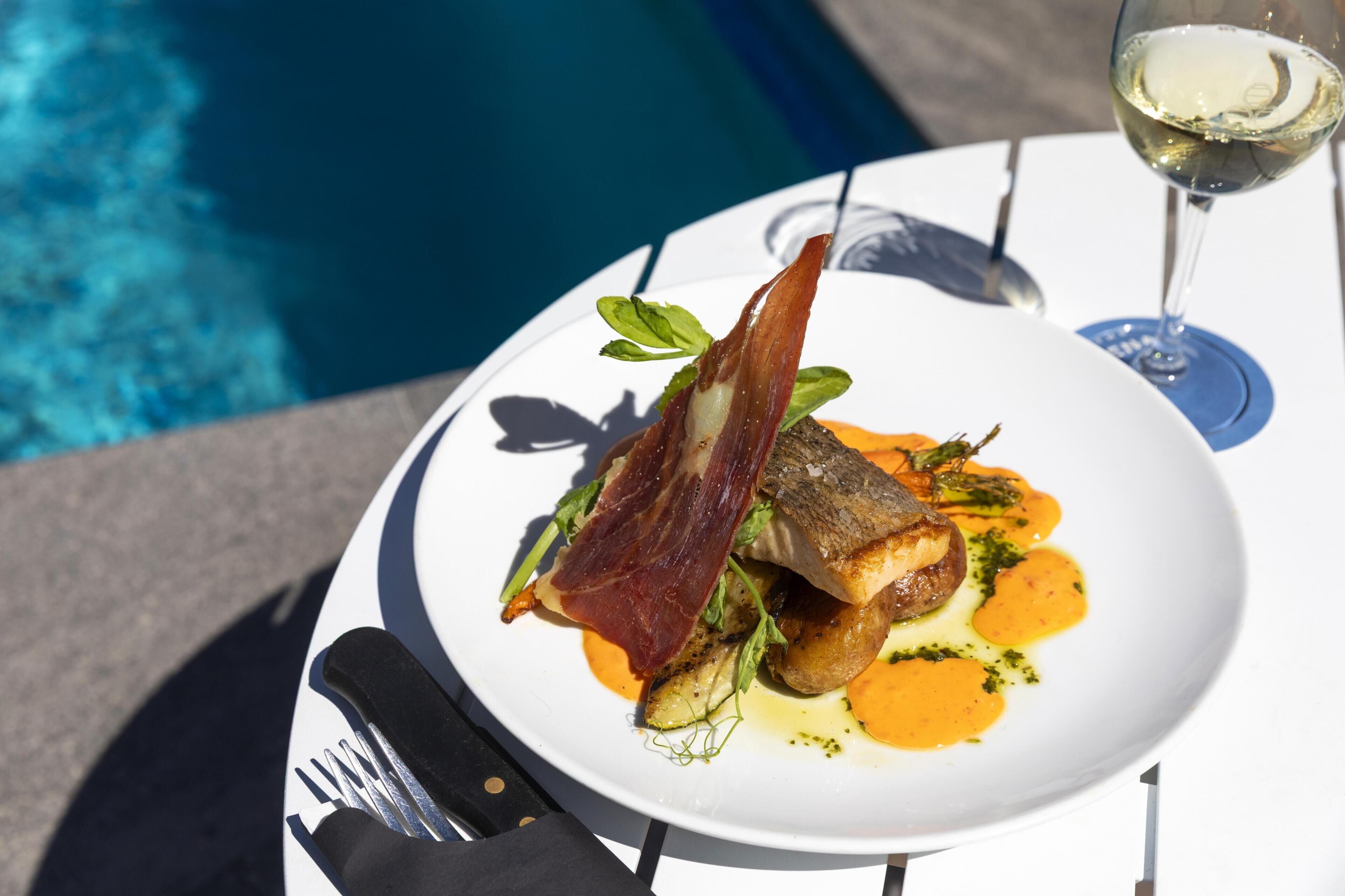 A dining table with a plate of food, a glass of white wine, and a pool in the background.