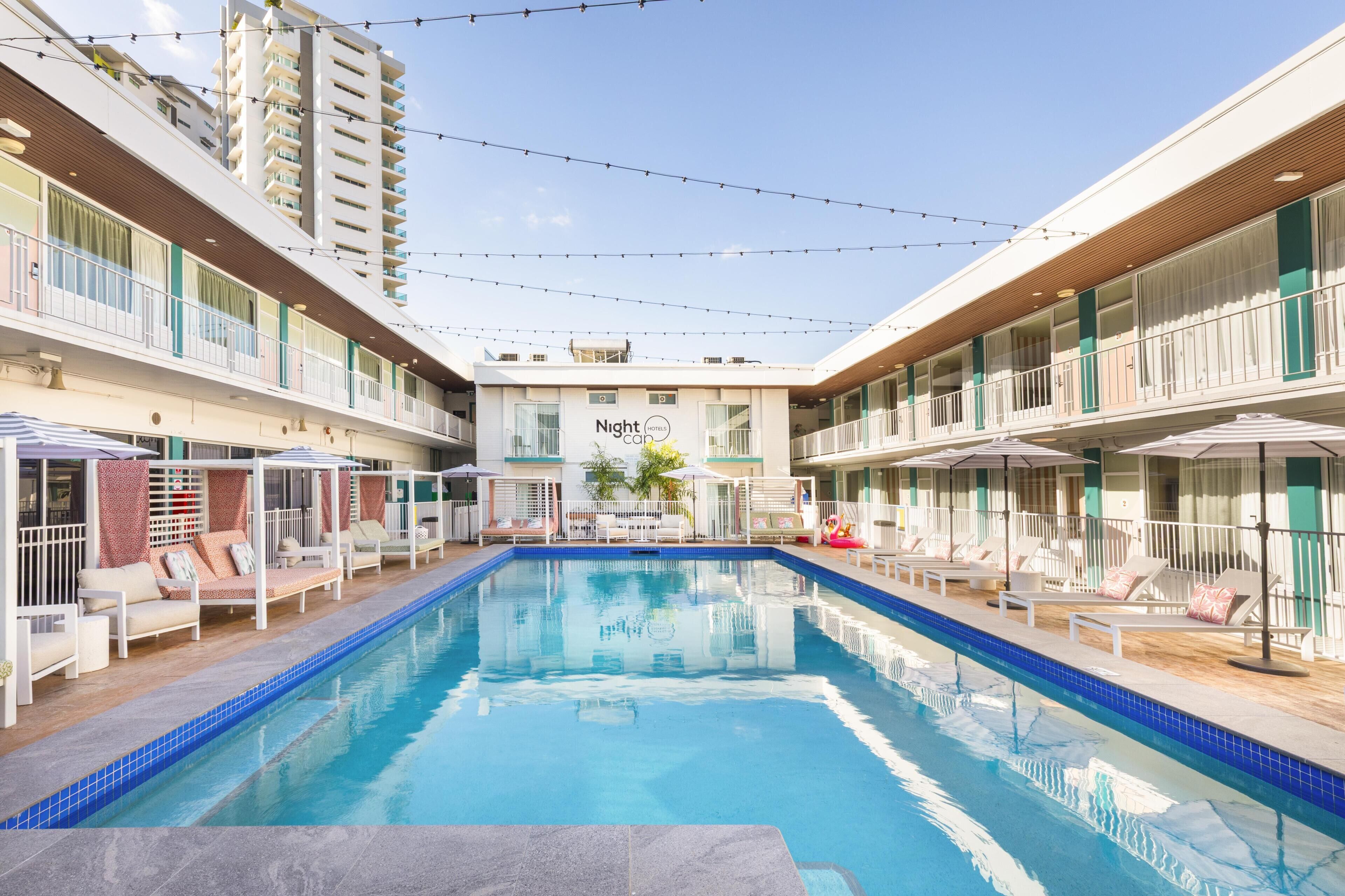 A swimming pool with lounge chairs and umbrellas, a building with balconies, and string lights.