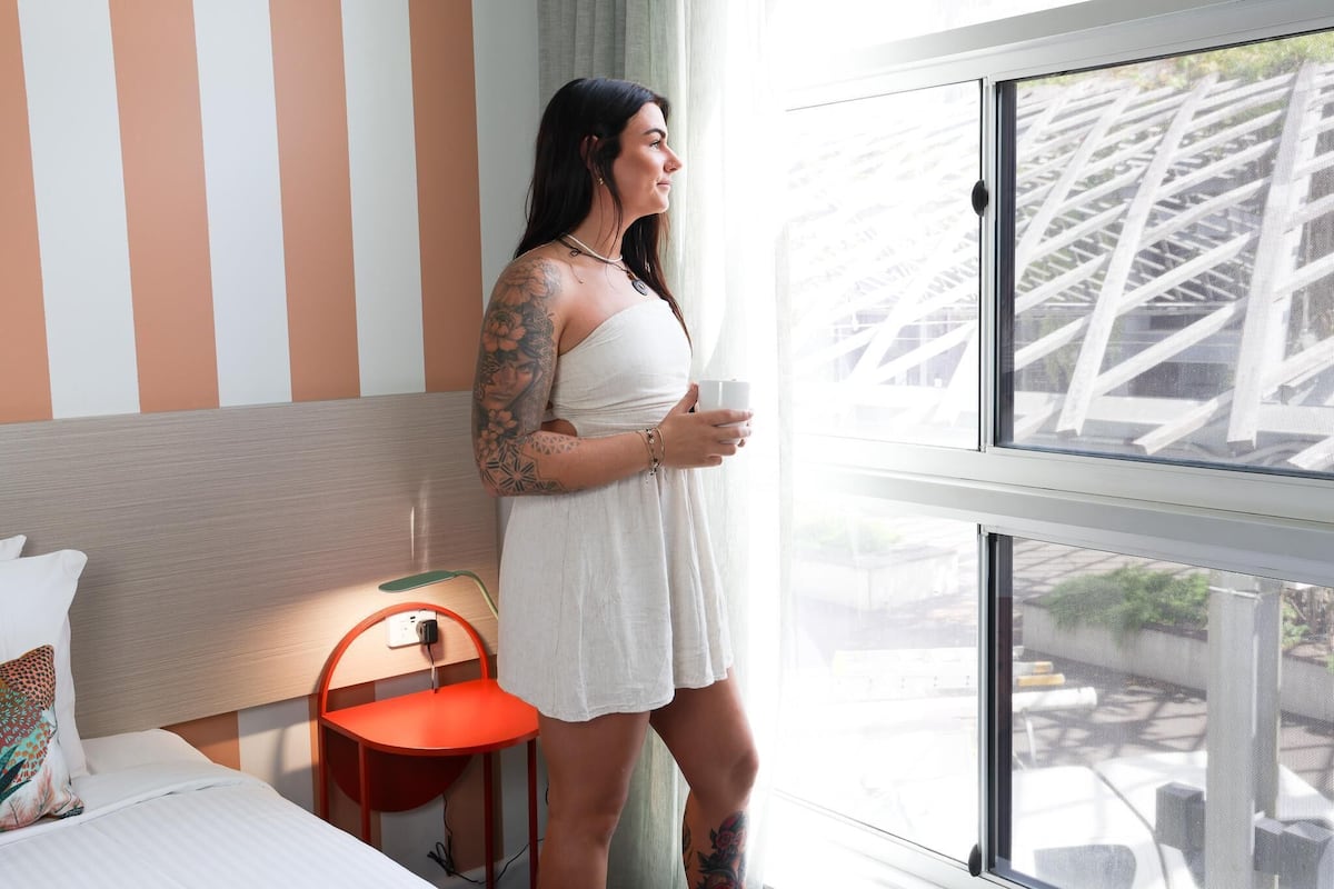 A person standing by a window in a room with striped walls, a bed, and a chair.