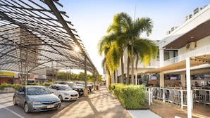 A covered outdoor area with a row of parked cars, a palm tree, and a building with outdoor seating. - Nightcap at the Cavenagh (Darwin)