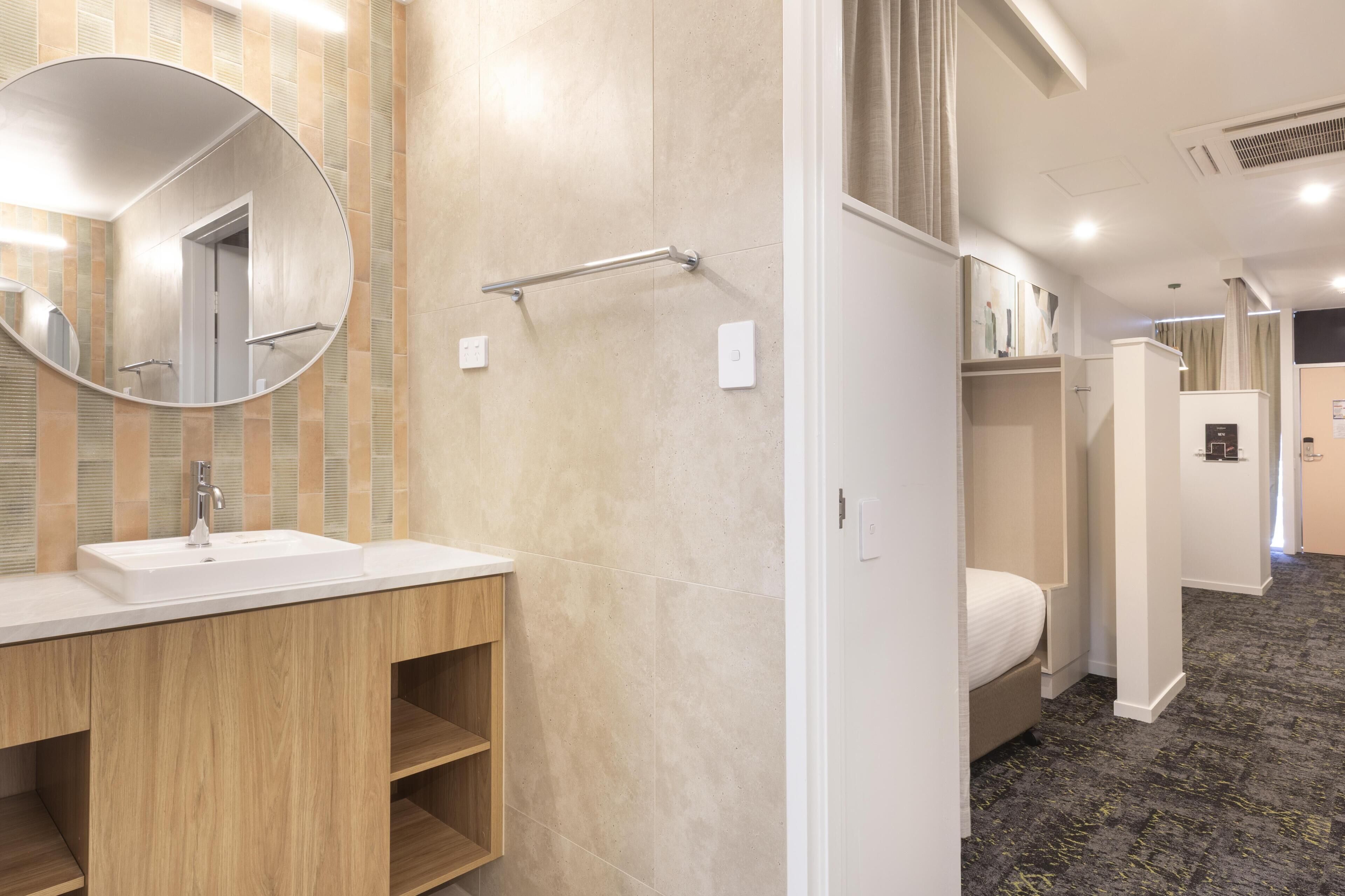 A hotel bathroom with a sink, mirror, towel rack, and a bed in the adjacent room.