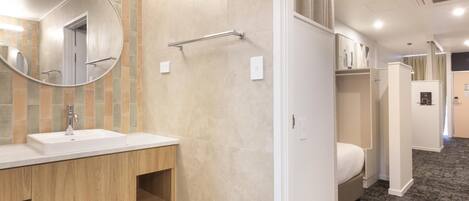 A hotel bathroom with a sink, mirror, towel rack, and a bed in the adjacent room.