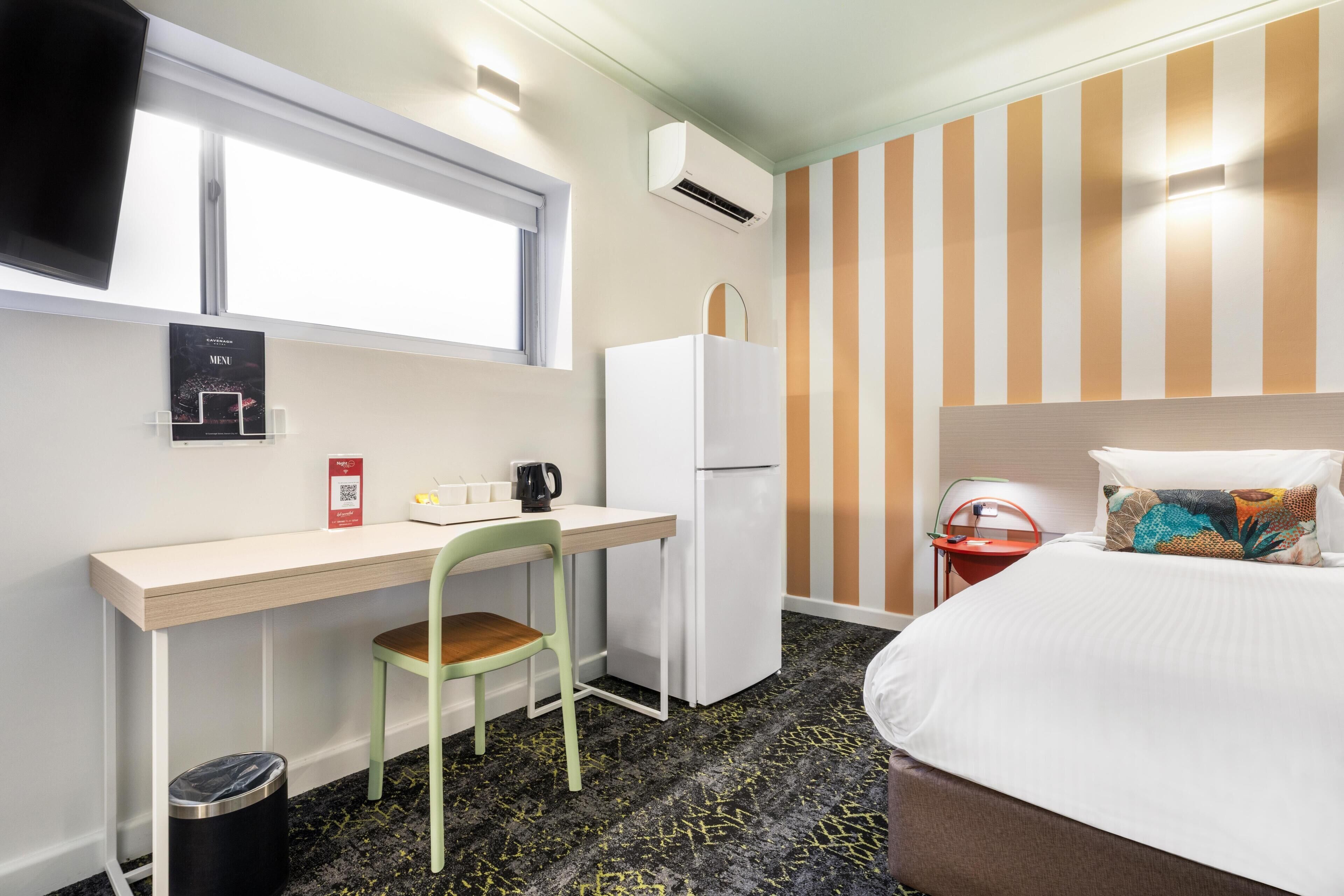 A hotel room with a bed, a desk with a chair, a refrigerator, a trash bin, a window, and a striped wall.