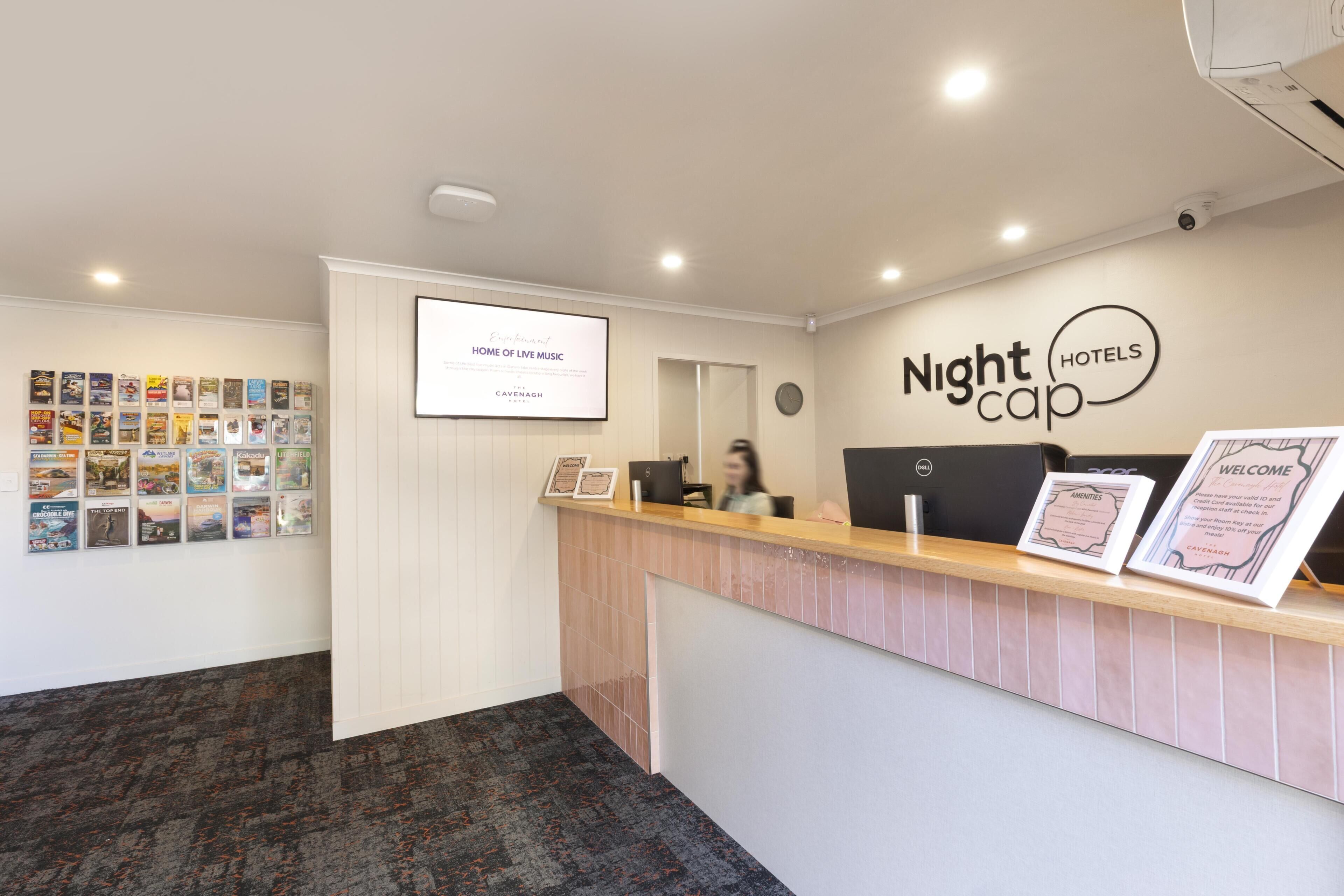 A hotel reception area with a reception desk, a wall-mounted screen displaying 'Entertainment Home of Live Music', and a wall with a 'Nightcap Hotels' sign.