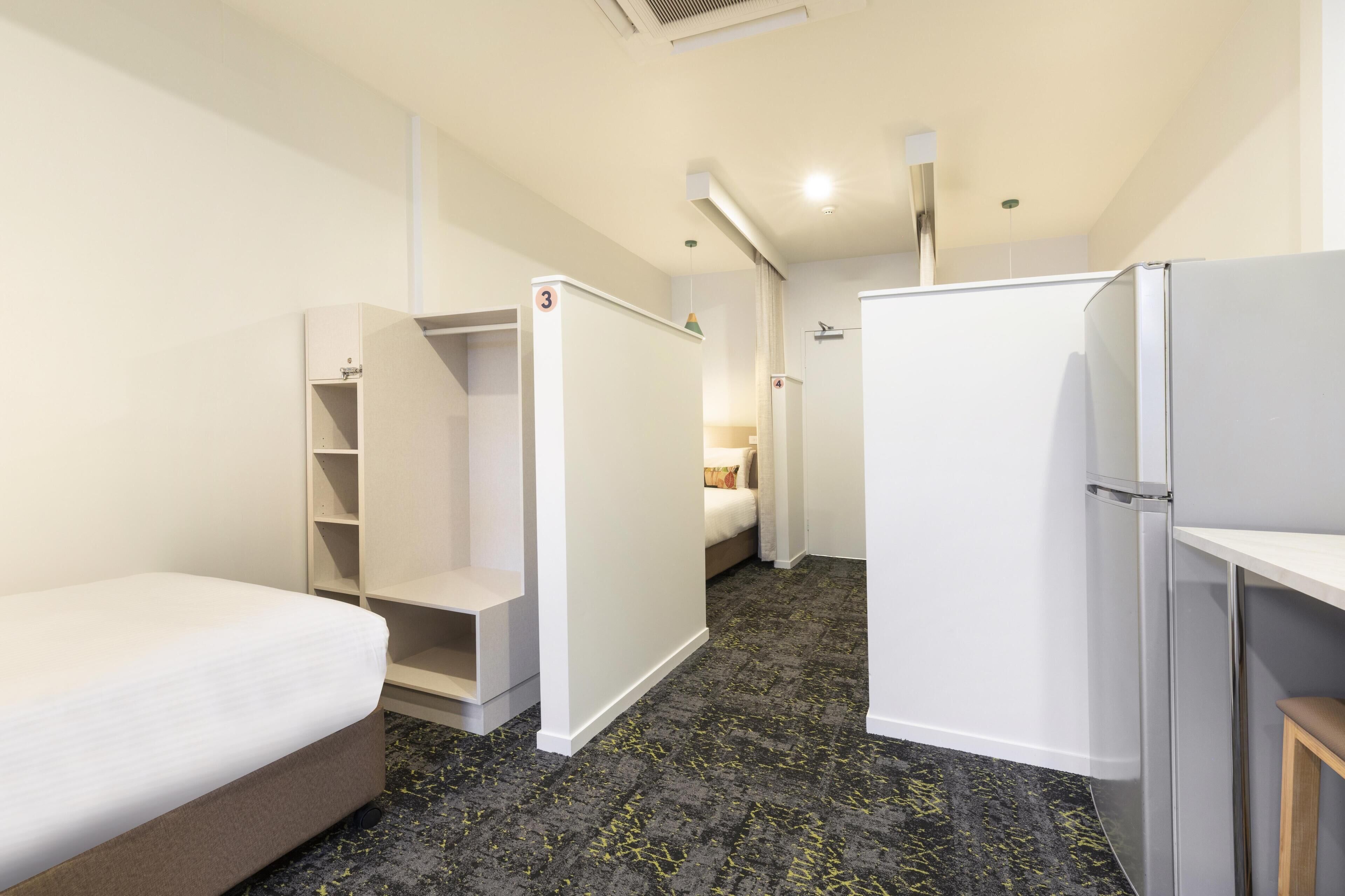 A compact hotel room with a bed, a wardrobe, a small desk, and a refrigerator.