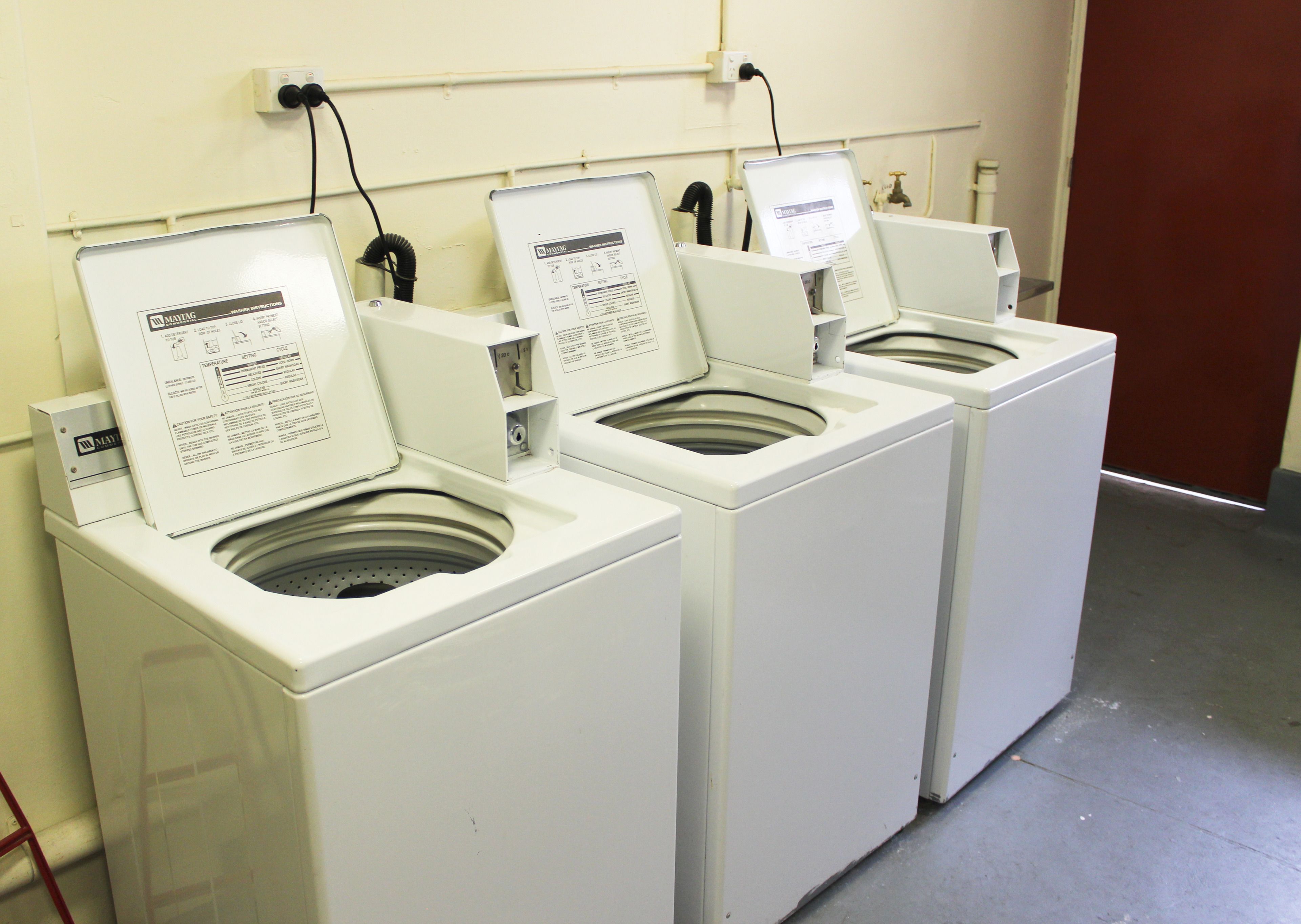 A row of white front-loading washing machines with instruction manuals open, connected to a power outlet on the wall.