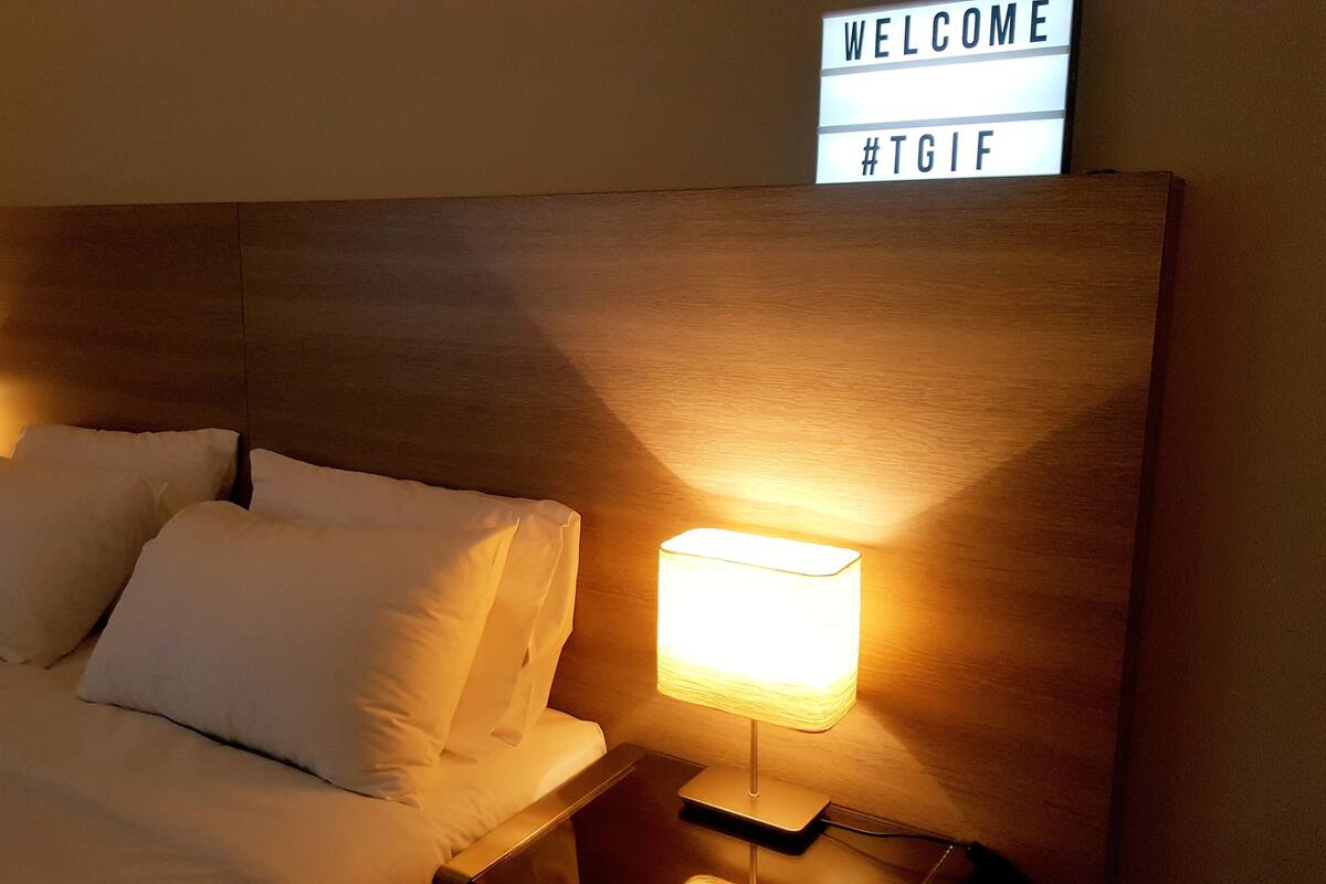 Quarto de hotel com cama arrumada, duas luminárias de cabeceira e um letreiro com a palavra 'WELCOME' e uma hashtag.