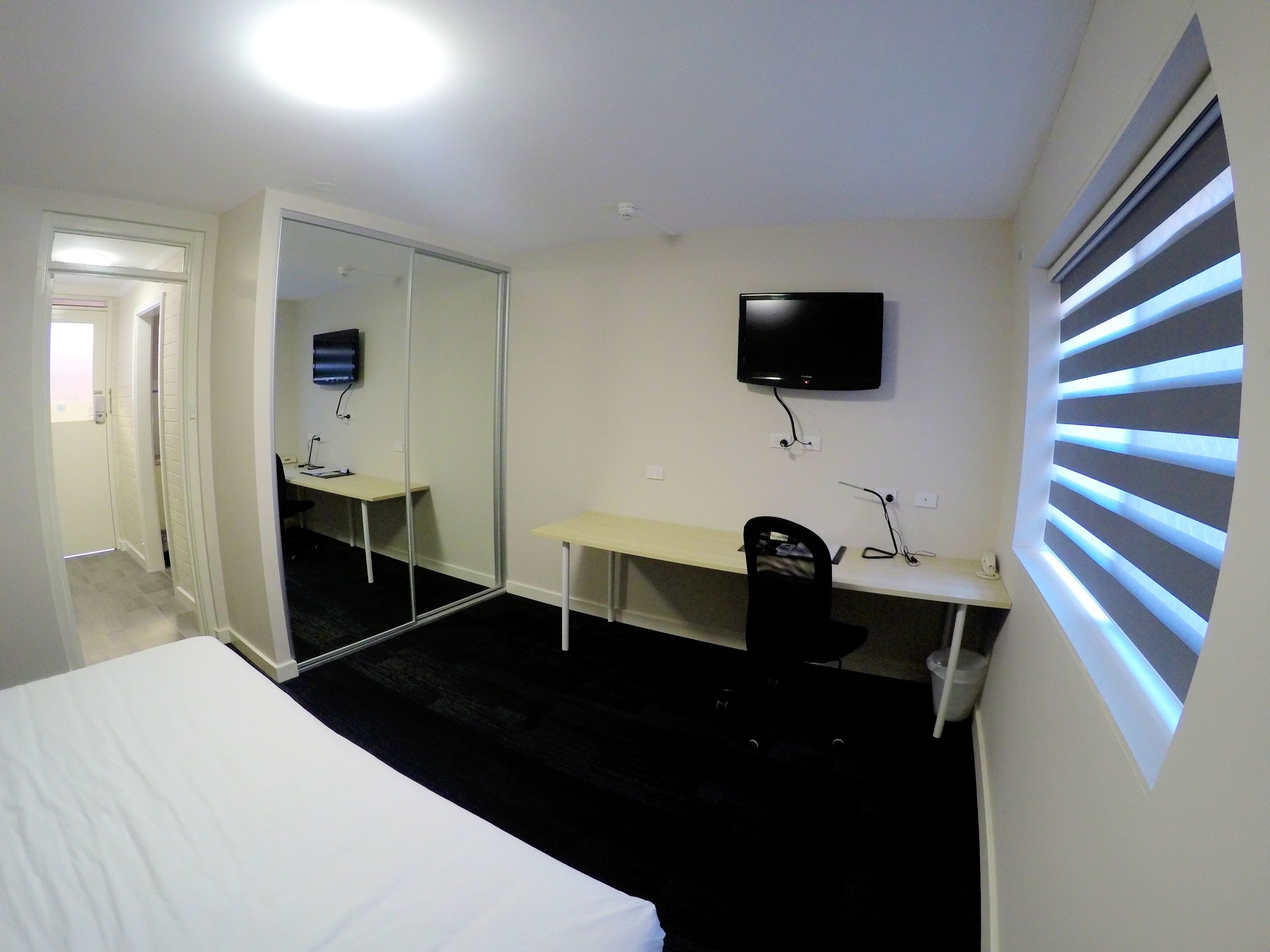 Habitación de hotel con cama, escritorio, silla, televisor y ventana con persianas.