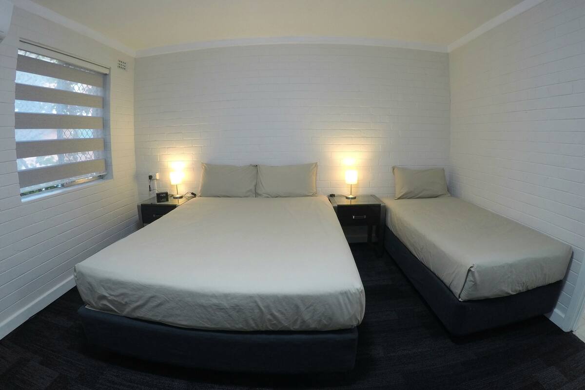 Quarto com duas camas, janela com persianas e luminárias de cabeceira.
