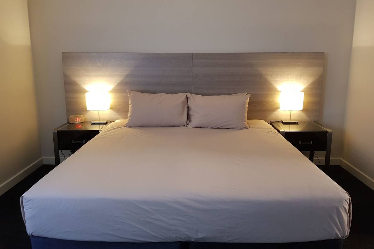 Uma cama bem arrumada com duas almofadas, duas mesas de cabeceira com abajures e uma cabeceira de madeira.