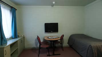 A small, single-room accommodation with a bed, a desk with a chair, a television, and a window with curtains.