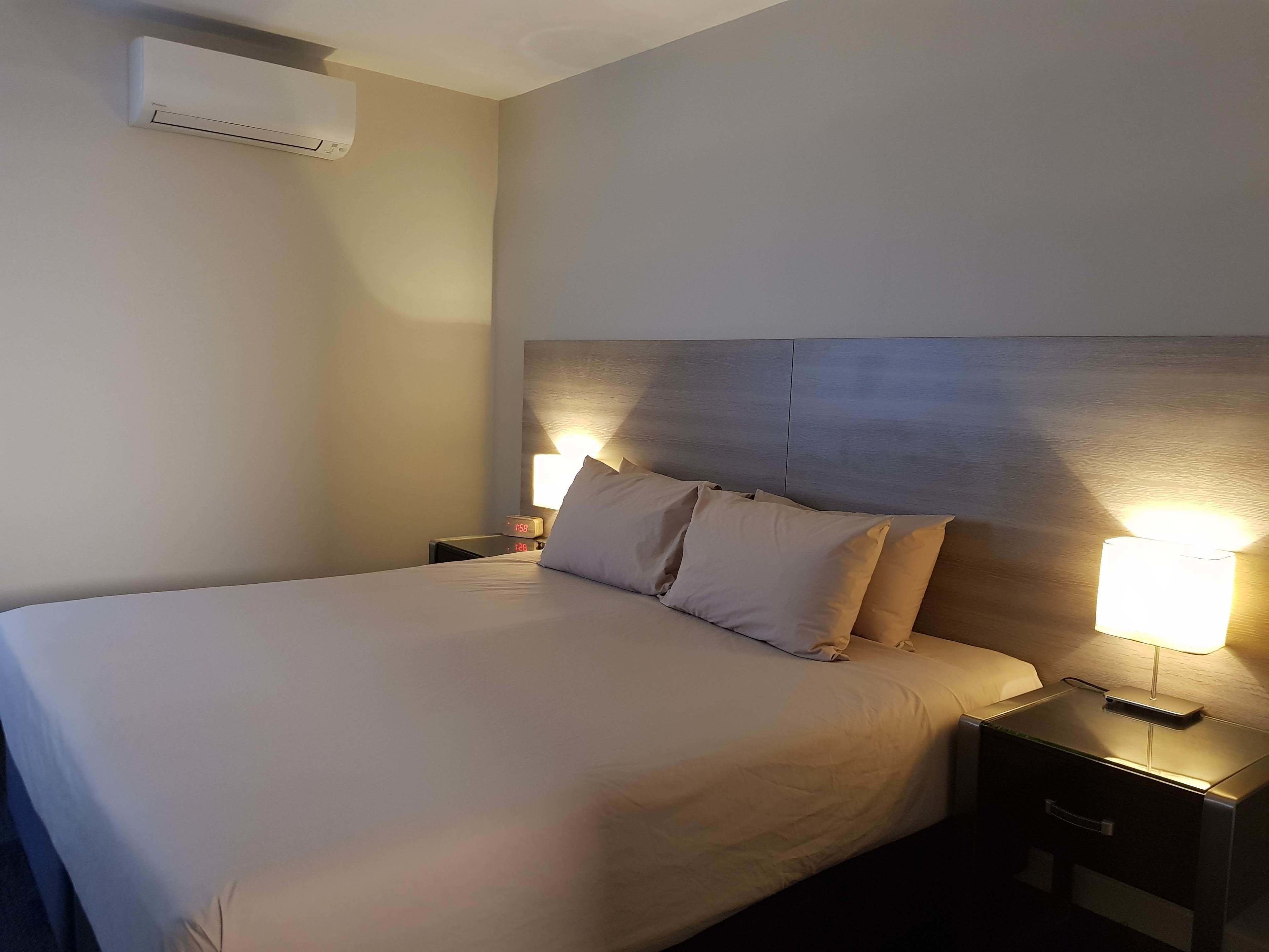 A hotel room with a bed, nightstands, a lamp, and an air conditioning unit.